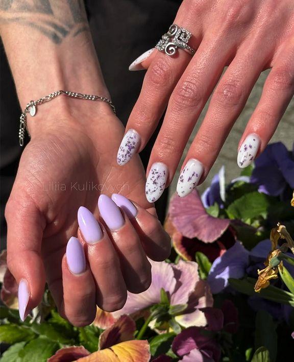 To hender med lange negler, en med lys lilla neglelakk og den andre med hvit neglelakk med blomsterdesign.
