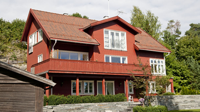 Rødt hus med balkong og store vinduer, omgitt av grønt landskap og trær.