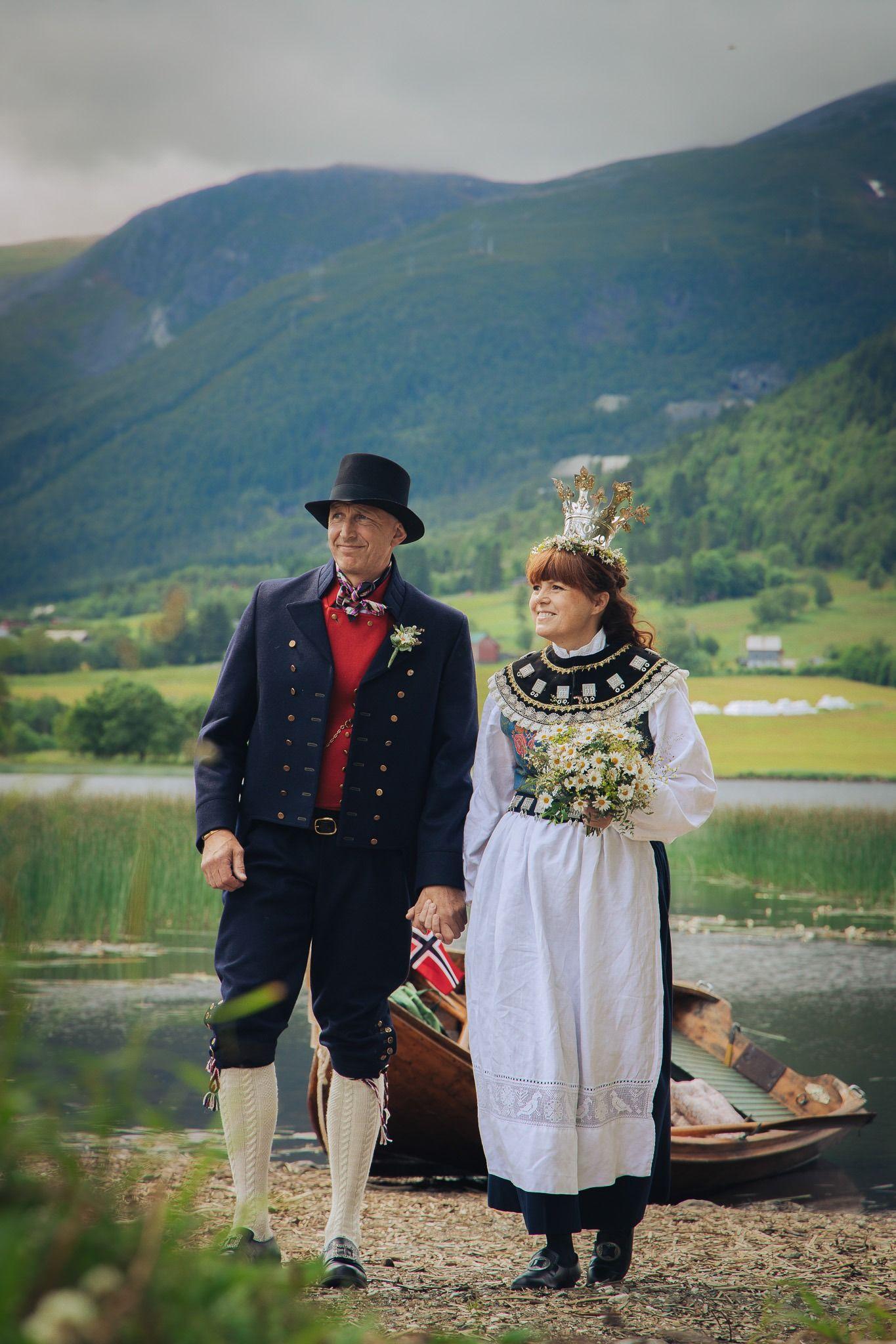 Kvinnelig og mannlig par i tradisjonelle norske bunader, holder hender ved vannkanten med fjell i bakgrunnen.