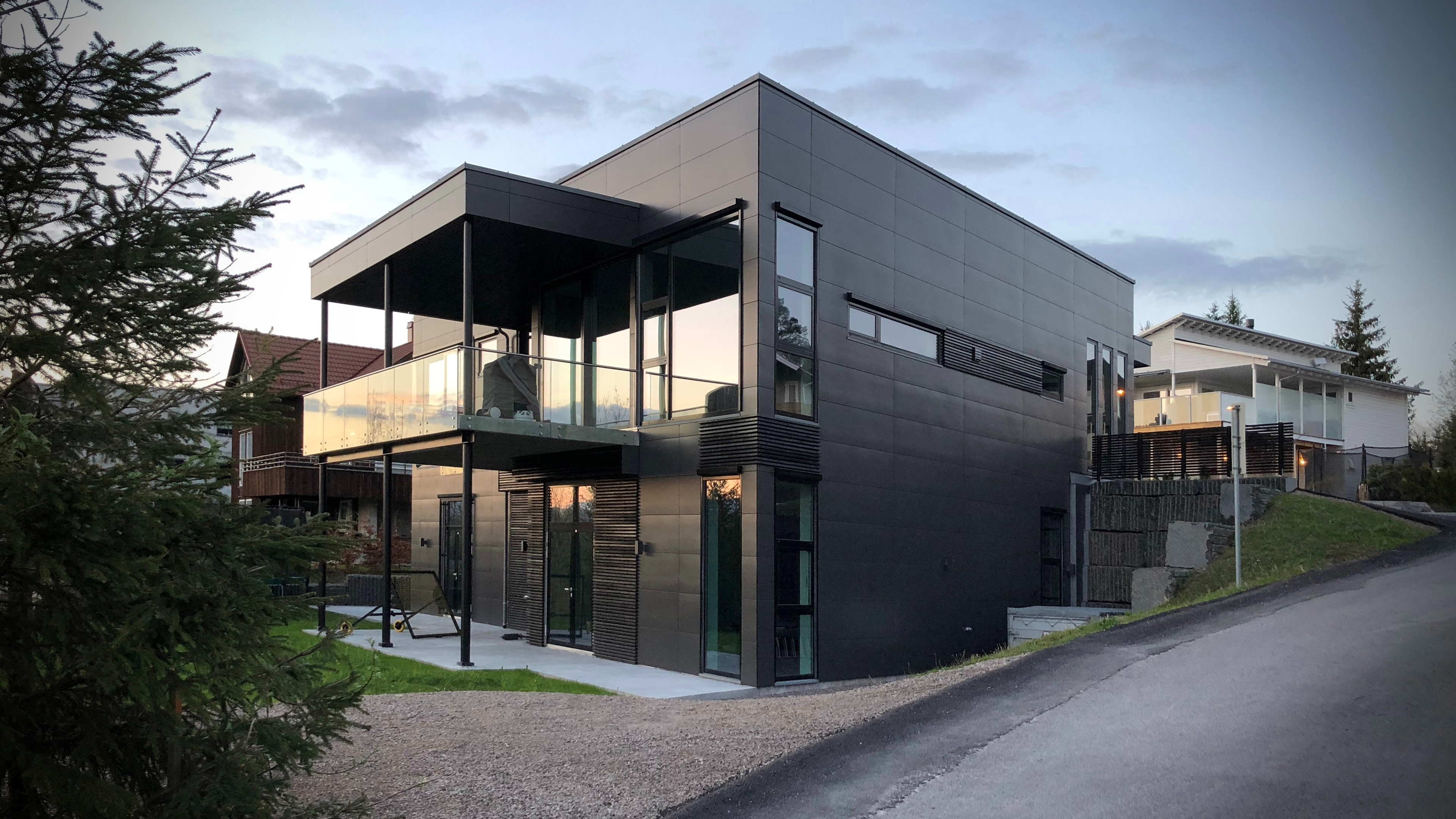 Moderne svart hus med store vinduer og balkong, beliggende på en skråning i et boligområde.