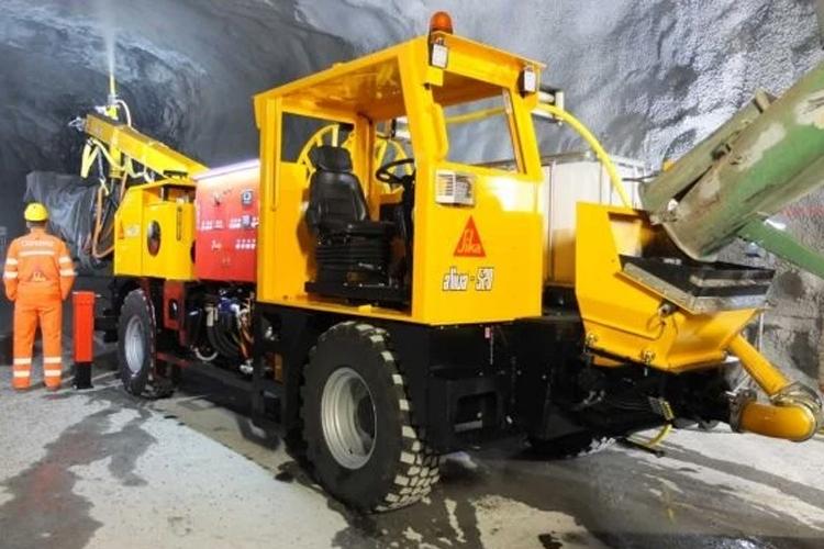 En stor gul tunnelmaskin med en sjåfør, plassert i en gruve med steinvegger. Arbeidere i oransje sikkerhetsklær er til stede.