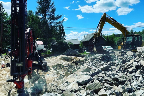 Gravemaskin og borerigg arbeider på en byggeplass med stein og grus under en blå himmel.