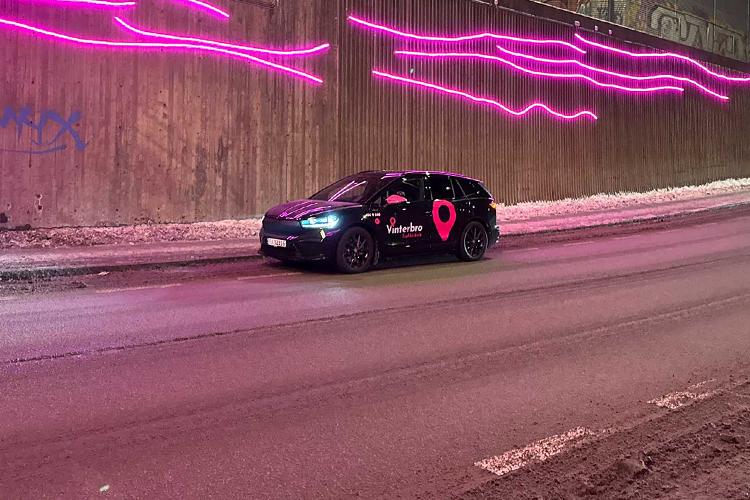 En svart bil med rosa detaljer står på en vei under en tunnel med rosa lyslinjer på veggen.