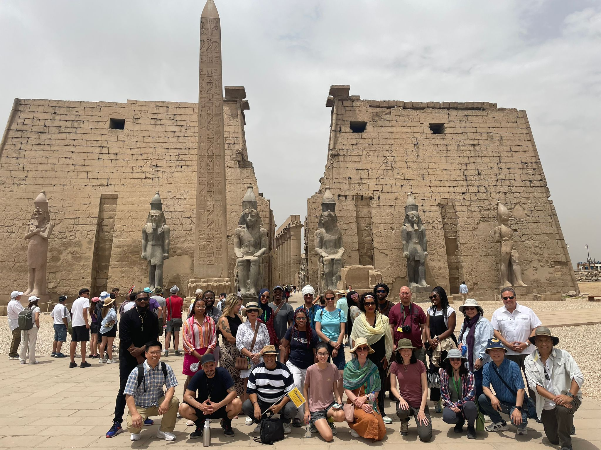En gruppe turister poserer foran de gamle templene i Luxor, med obelisker og statuer i bakgrunnen.