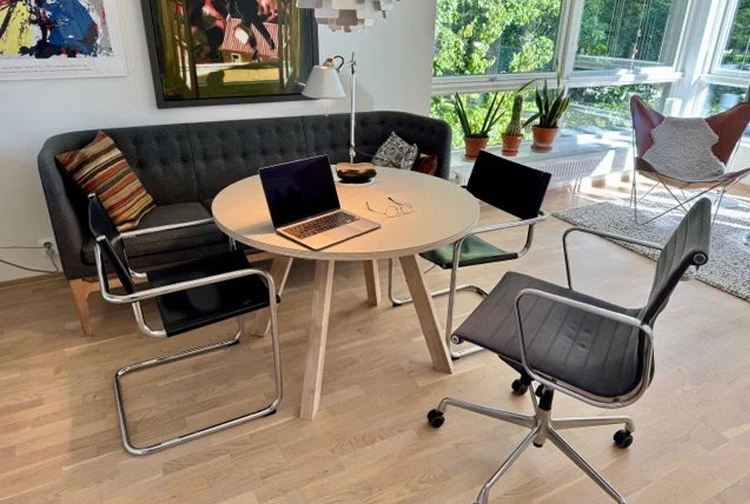 Moderne kontorinnredning med et rundt bord, to datamaskiner og stoler i et lyst rom med grønne planter.