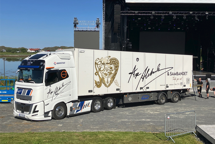 En stor lastebil med kunstnerens signatur parkert foran en scene, klar for en konsert.