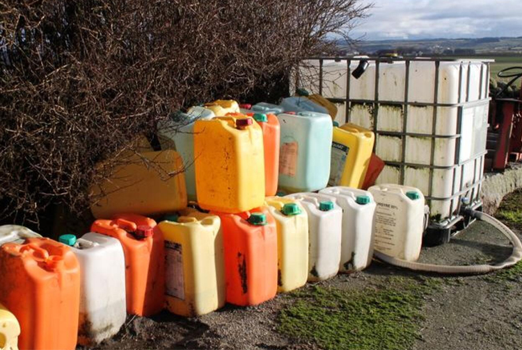 Stablede plastkanner i ulike farger, plassert ved en vegg med en stor tank i bakgrunnen.