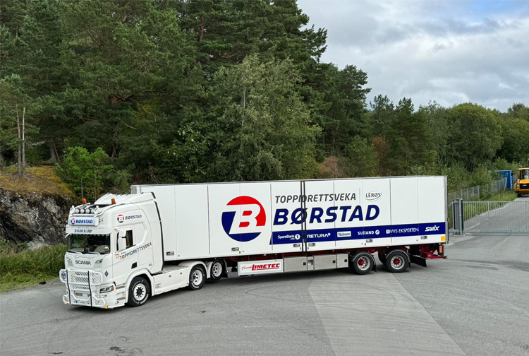 En lastebil med Børstad-logoen står parkert i et industriområde omringet av trær.