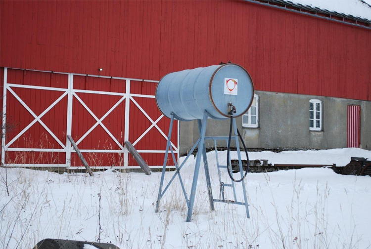 En blå vanntank på stativ foran en rød låve, omringet av snø og tørre gress.