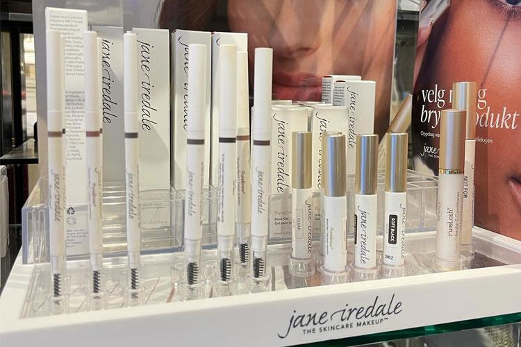 Utvalg av Jane Iredale sminkeprodukter på en hylle, inkludert leppestifter, øyebrynspenn og foundation.