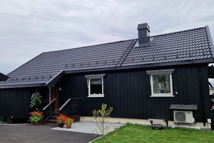 En svart trehytte med takstein, blomster i hagen og en liten terrasse foran inngangsdøren.