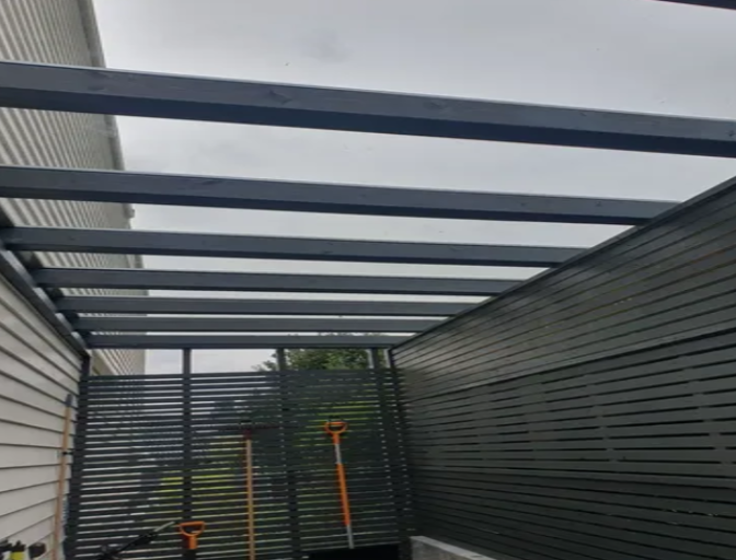 Takramme av metall med horisontale bjelker, delvis dekket av vegger, i en uteplass med hageredskaper.