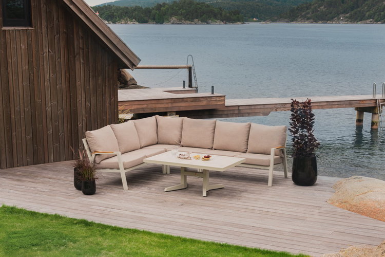 Uteplass med en moderne sofa og bord ved vannet, omkranset av grønt gress og en trebygning i bakgrunnen.