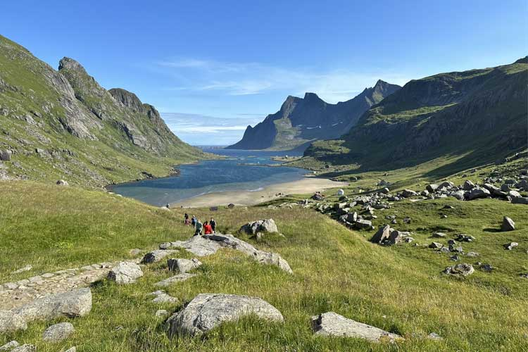 Fjellandskap med en dal, en innsjø og turister som går langs stien i solfylt vær.