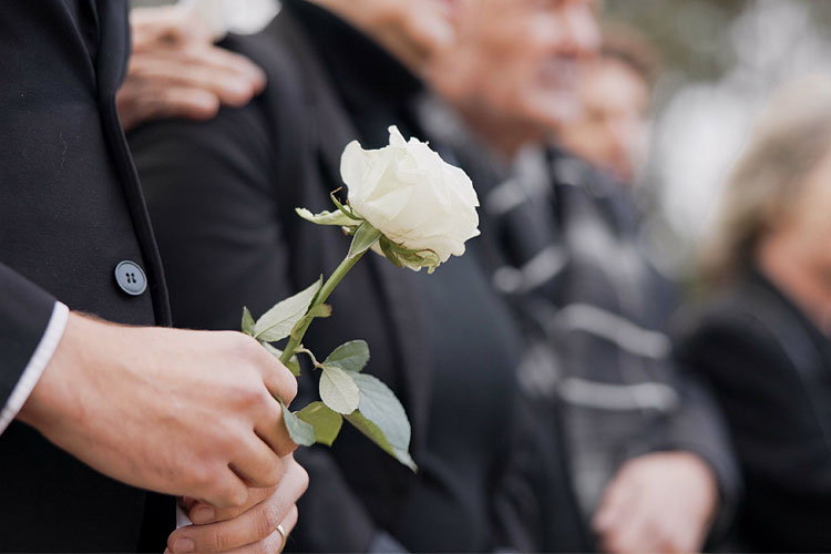En person holder en hvit rose, mens en gruppe mennesker står sammen i bakgrunnen under en seremoni.