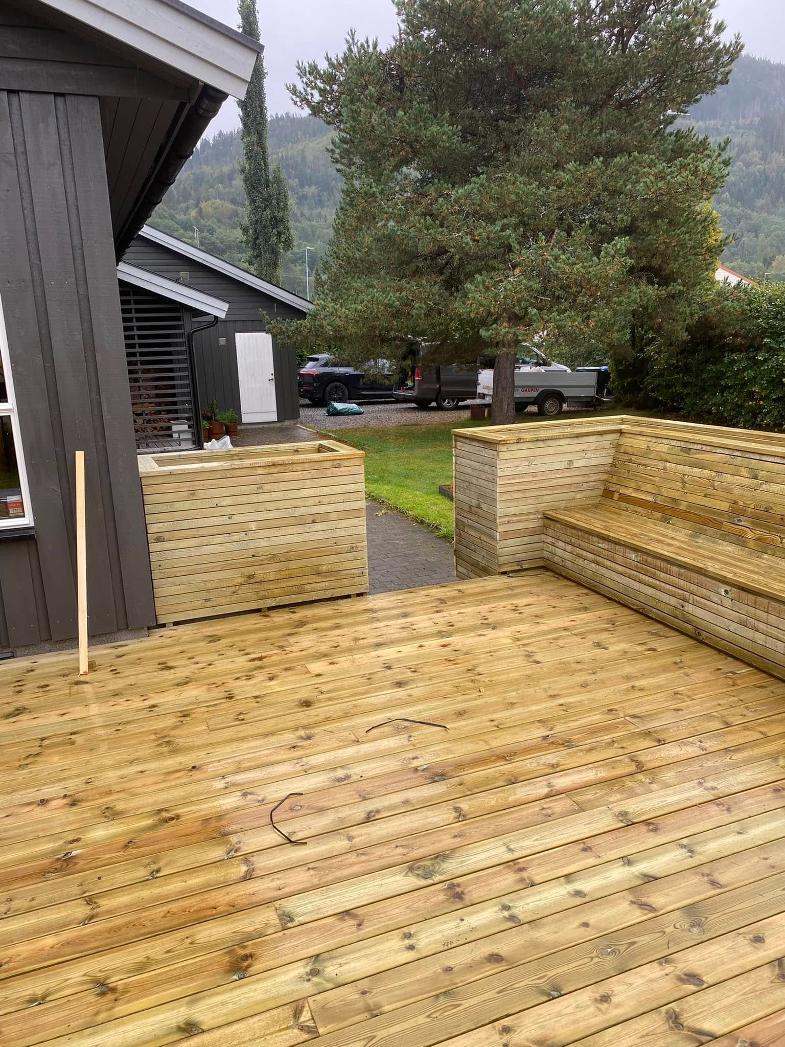 Terrasse med treverk, sitteplasser og planter, omgitt av grønt landskap og bygninger i bakgrunnen.