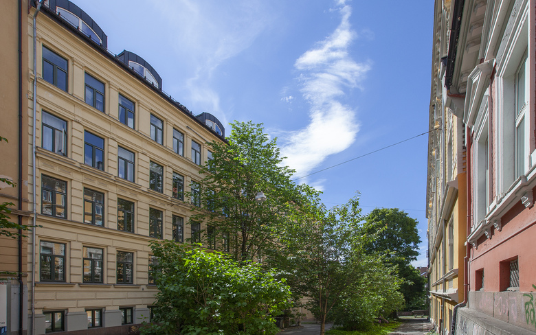 En solfylt gate med bygninger på hver side og et tre i midten, under en klar blå himmel med noen skyer.