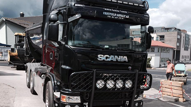 En svart Scania lastebil med lysbjelke, parkert på en gate med bygninger i bakgrunnen.