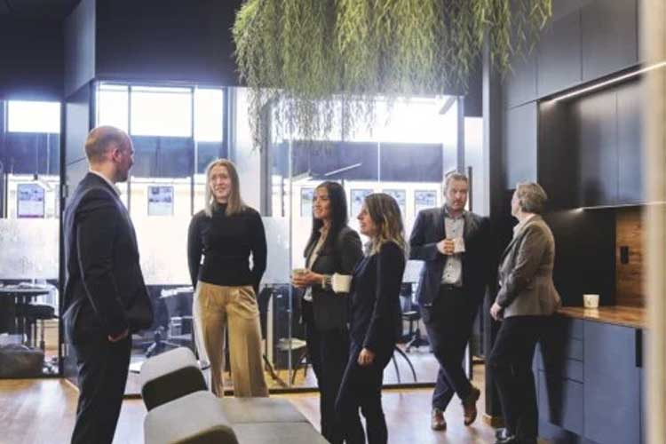 En gruppe mennesker i et moderne kontorlandskap, som diskuterer og drikker kaffe. Grønne planter henger fra taket.