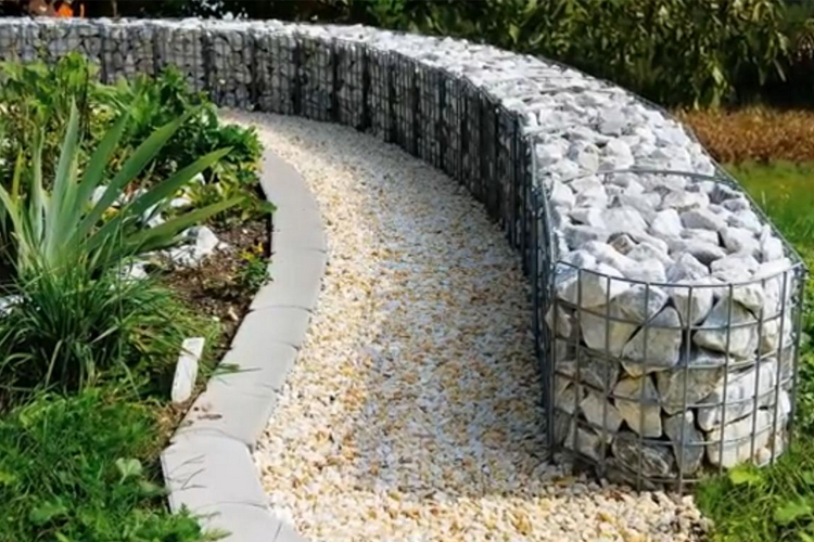En buet sti laget av småstein, omgitt av gitterkasser fylt med steiner og grønne planter.