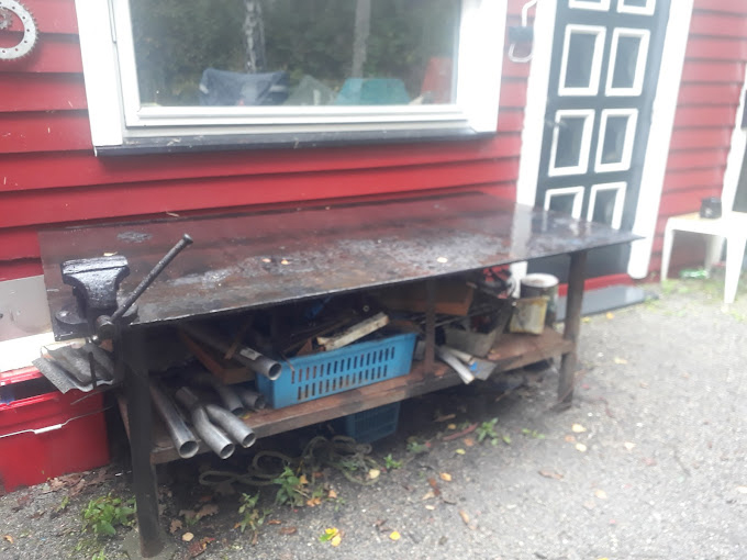 Et rustikt arbeidsbord med verktøy og materialer, plassert foran en rød husvegg.