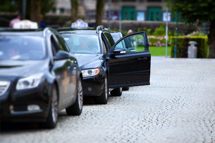Svart taxi med åpen dør parkert på brosteinsbelagt plass i en bypark.