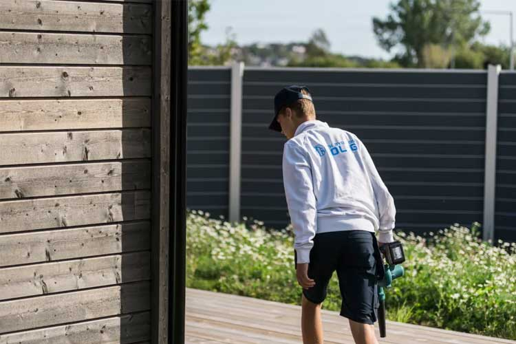 En ung gutt i hvit hettegenser og shorts, som går ut av en bygning med en gressklipper i hånden.