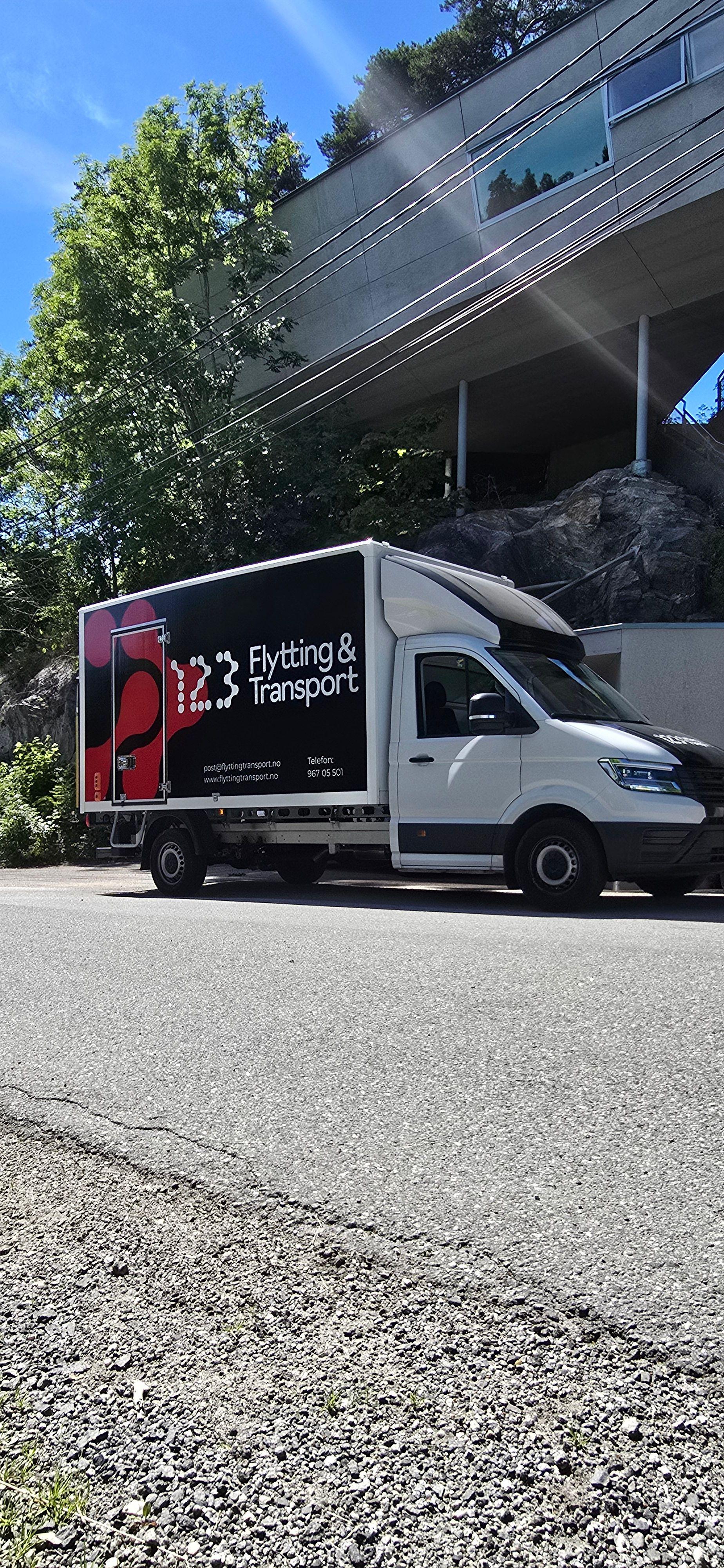 En hvit lastebil med logoen til 123 Transport parkert langs veien, med trær og bygninger i bakgrunnen.