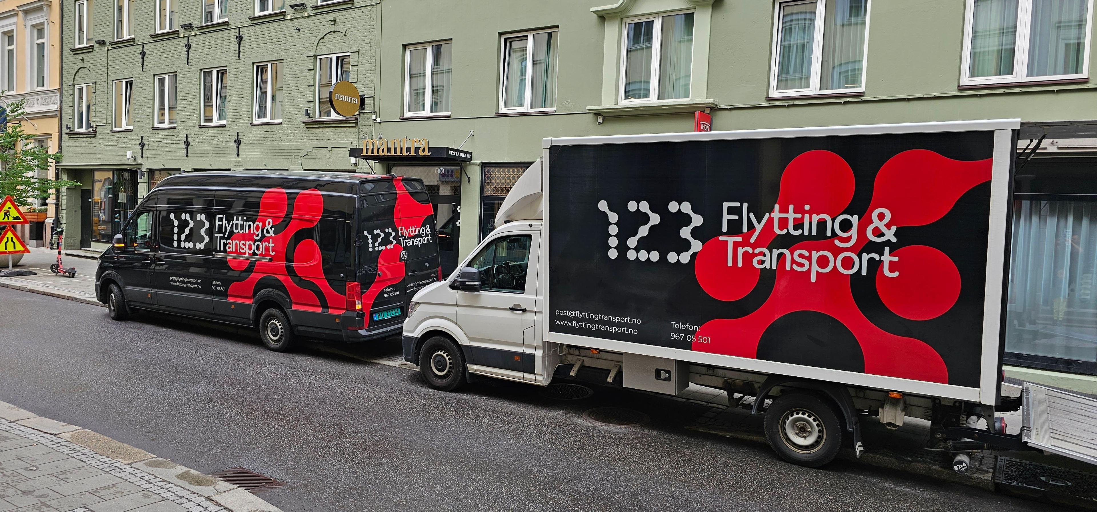 To lastebiler med logoen til 123 Flytting & Transport parkert på en gate med bygninger i bakgrunnen.