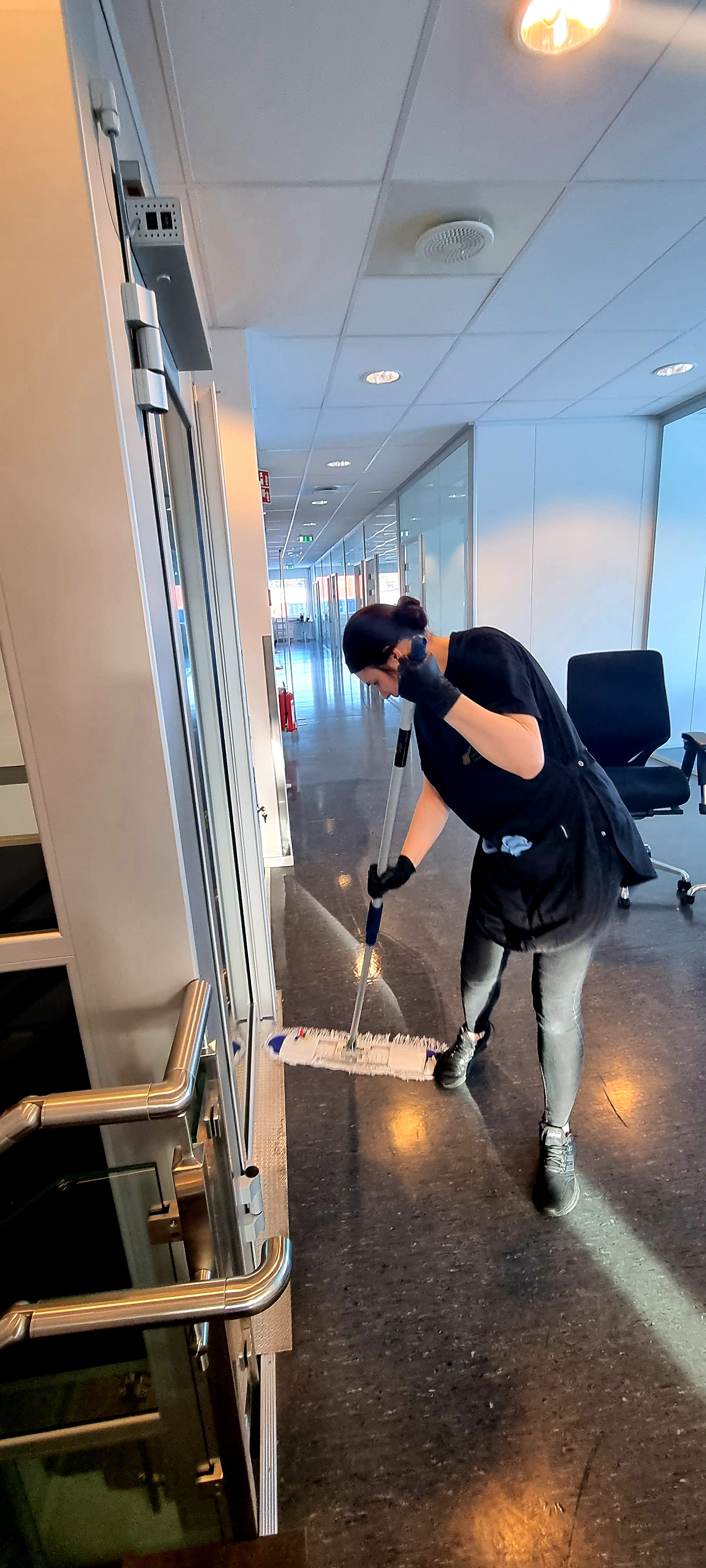 En kvinne rengjør en dør med en mopp i et moderne kontorlandskap.