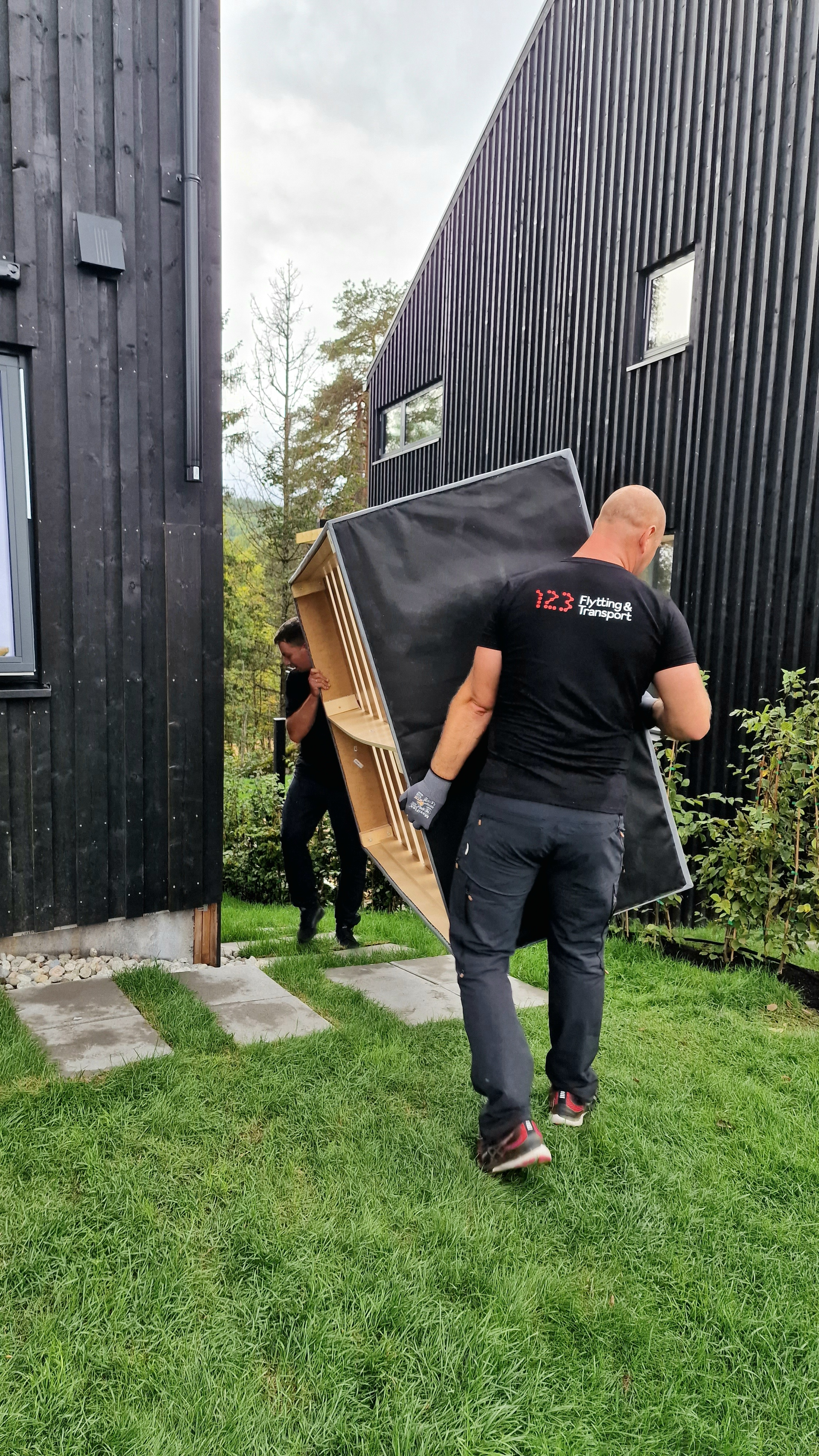 To personer bærer en stor svart sofa ut av en moderne hytte i hagen.