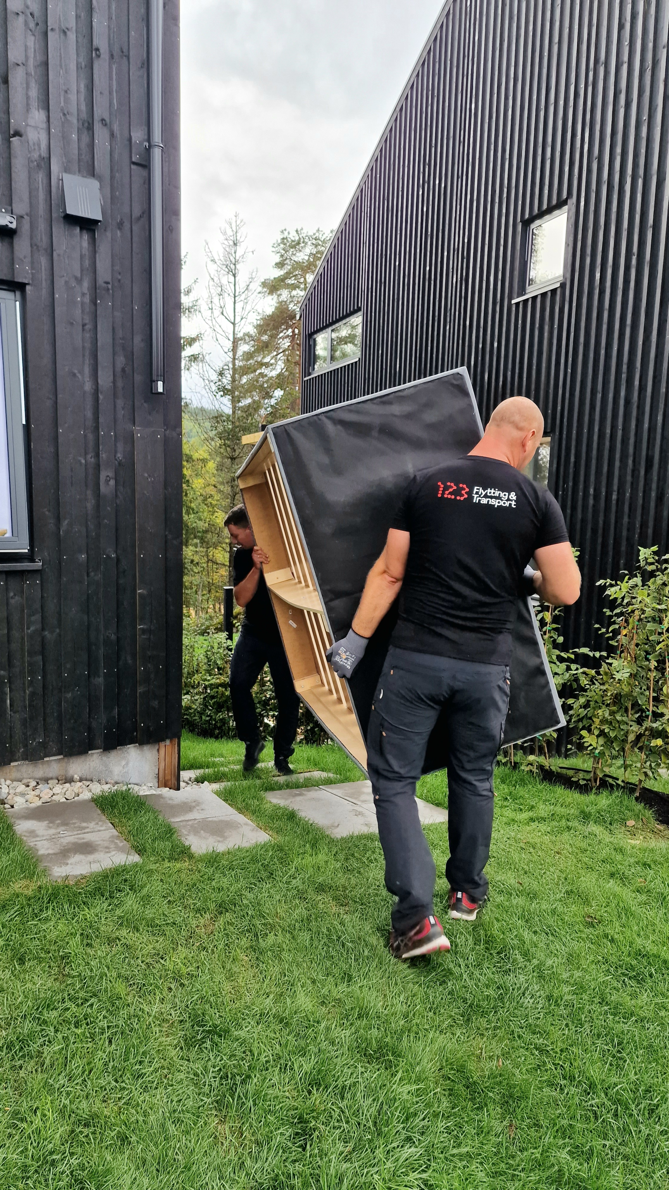 To personer bærer en stor svart sofa ut av en moderne hytte i hagen.