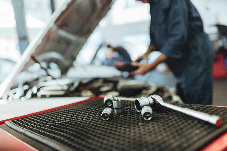 Verktøysett med skralle og muttere på en arbeidsbenk i et bilverksted. Mekaniker jobber med motoren i bakgrunnen.
