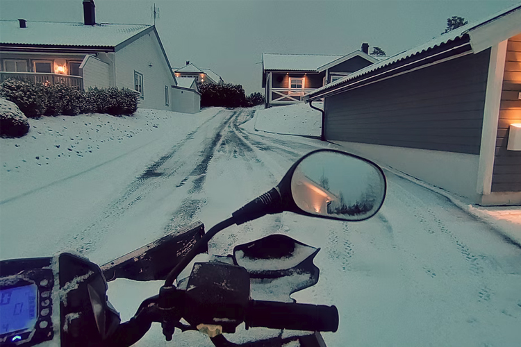Snølagt vei med hus i bakgrunnen, sett fra en snøskuter med speil og instrumentpanel i forgrunnen.
