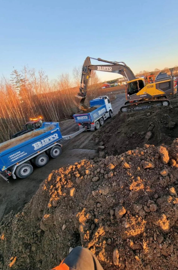 Gravemaskin som arbeider på en byggeplass med lastebil og jordhauger i bakgrunnen under soloppgang.