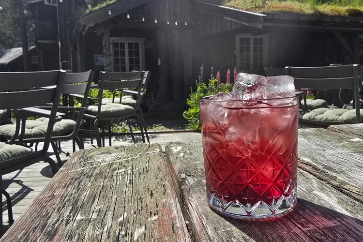 En glass med rød drink med isbiter, plassert på et gammelt trebord, med en hytte i bakgrunnen.