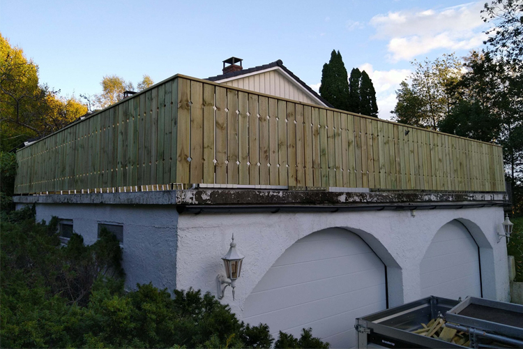 Terrasse med trerekkverk på taket av en garasje, omgitt av trær og en blå himmel.