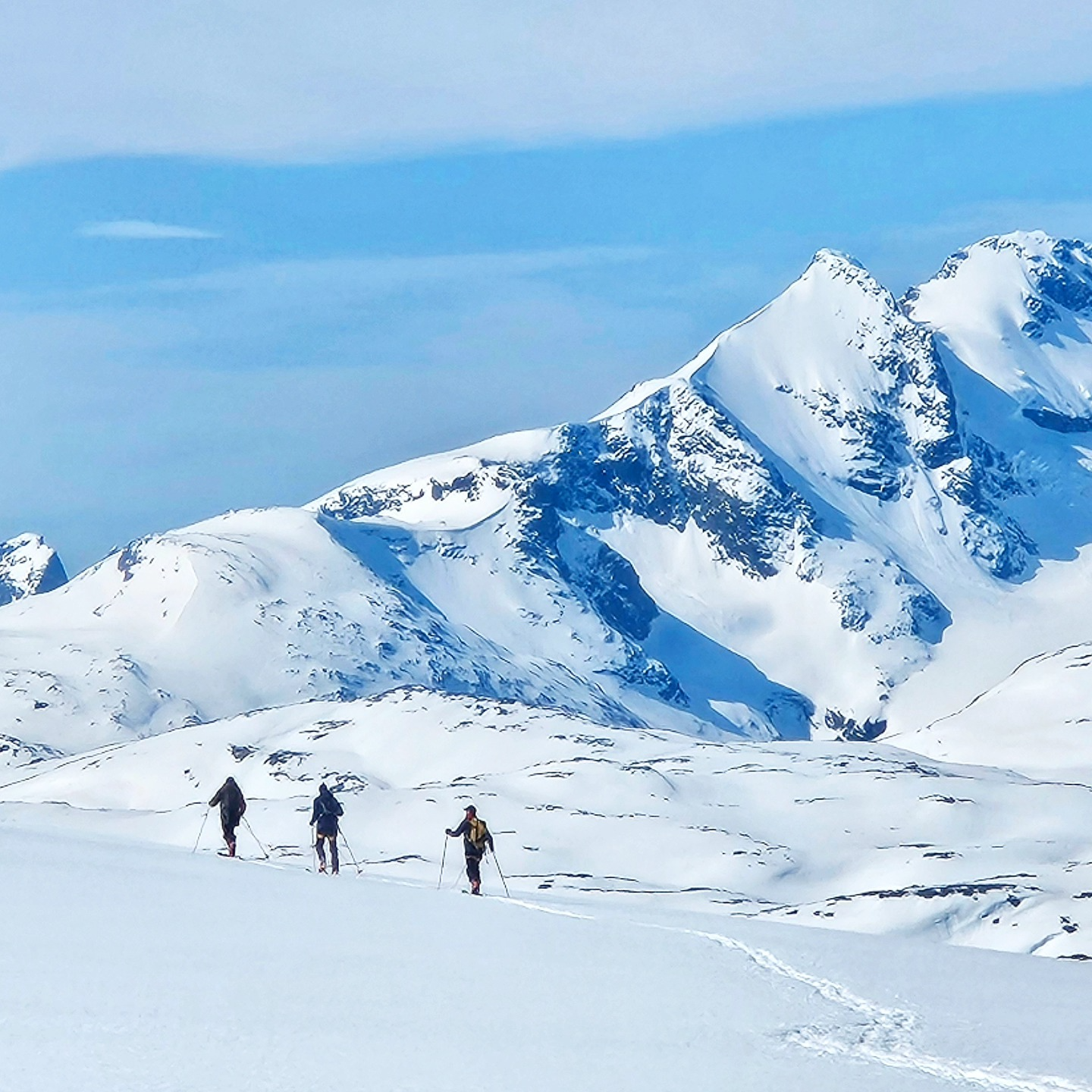 Tre personer går på langrenn i snødekte fjell med blå himmel i bakgrunnen.