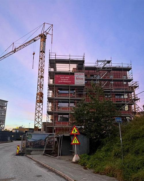 Bygget med stillas og kran, under konstruksjon, med veiskilt og grønt område i forgrunnen.