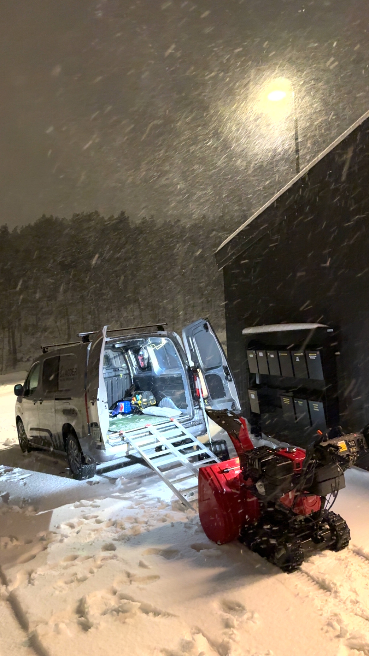 En snøfreser står ved siden av en bil med åpen bakluke, snøfall og mørk bakgrunn.