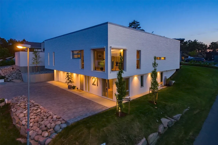 Moderne hus med store vinduer, beliggende i et rolig nabolag, opplyst av gatebelysning ved skumring.