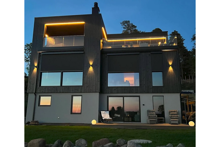 Moderne hus med store vinduer og balkong, opplyst av lys i skumringen ved sjøen.