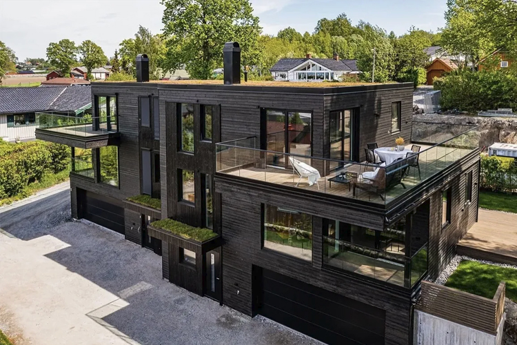 Moderne svart hus med store vinduer og terrasser, omgitt av grønt landskap og nabolag.