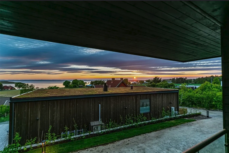 Utsikt fra en terrasse med solnedgang over havet og grønne hager i forgrunnen.