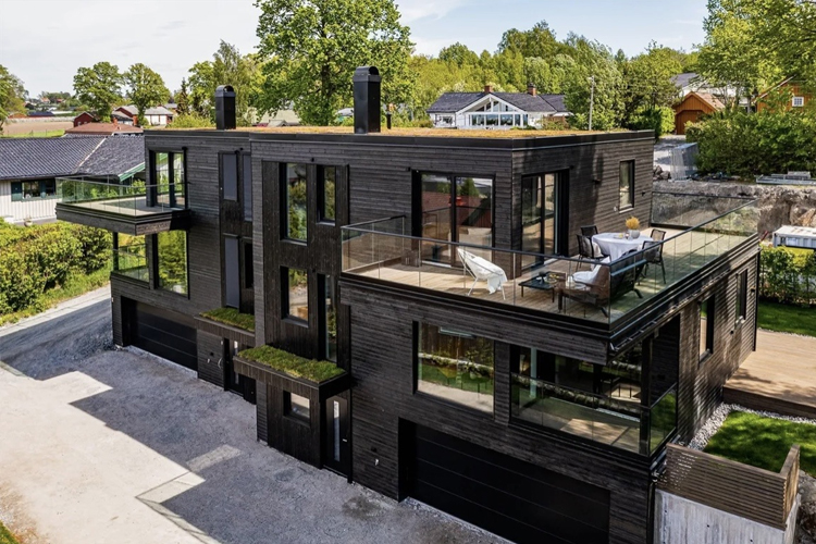 Moderne svart hus med store vinduer og balkonger, omgitt av grønt landskap og nabolag.