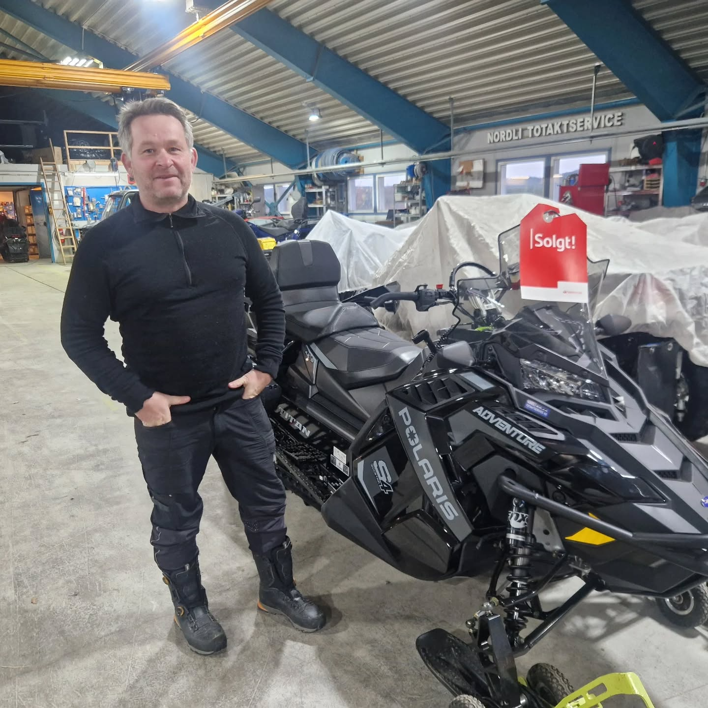 Mann står ved en svart snøscooter i et verksted, med "Solgt" skilt på scooteren.