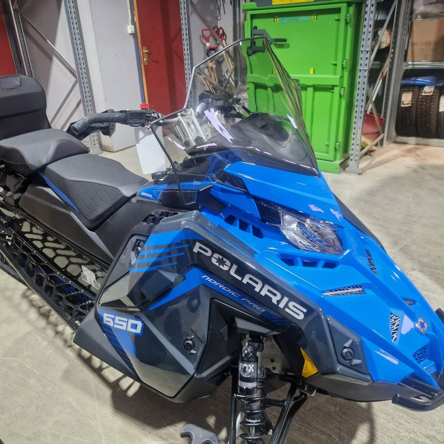 En blå snøscooter av merket Polaris 650, parkert i et verksted med verktøy og hyller i bakgrunnen.