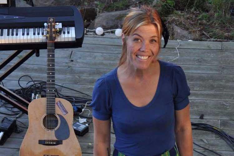 Kvinne med smilende ansikt står foran en akustisk gitar og et keyboard på en utendørs scene.