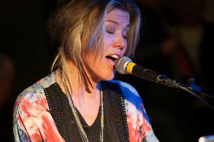 Kvinne synger med mikrofon mens hun spiller piano, iført en fargerik topp.