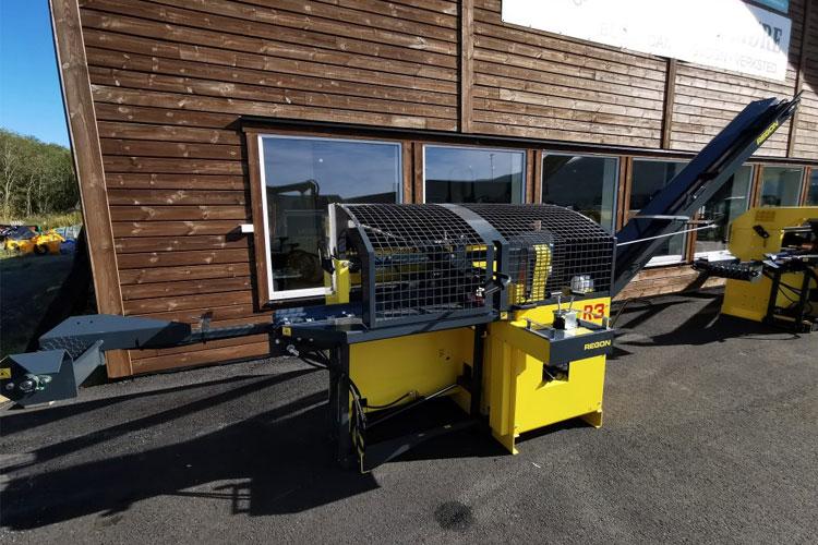 En gul og svart trebearbeidingsmaskin med transportbånd, plassert foran en bygning med vinduer.